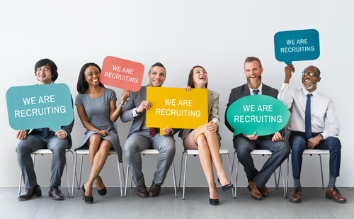 A group of business professionals seated with signs reading "We Are Recruiting" to attract potential job applicants.